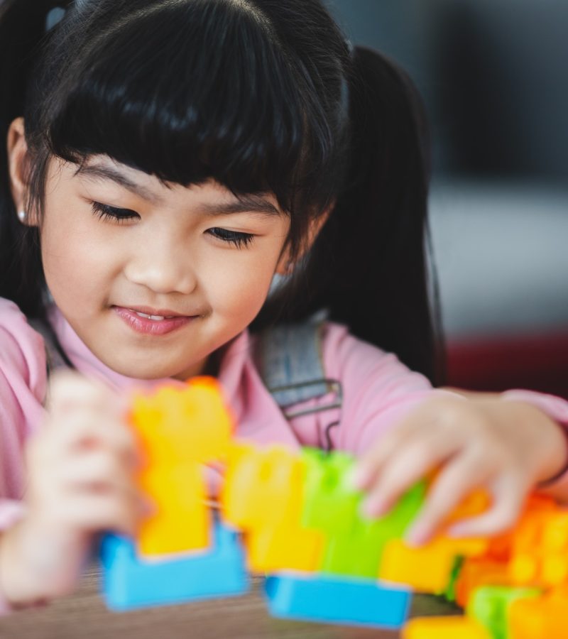 Cute little Asian girl playing alone with lego game blocks on table trying to build